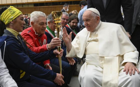 Pope Francis, sitting, blesses a gold staff held by a man and a woman
