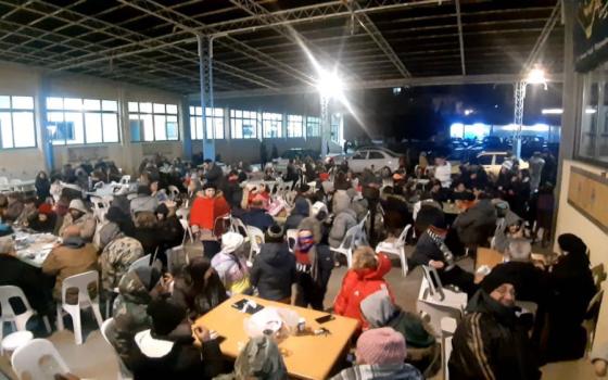 Many people in winter clothing gather at tables in a warehouse-looking space