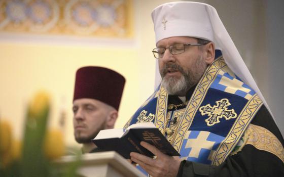 An older white man with glasses wears a round circular hat with a veil and a blue and gold stole with crosses on it. He reads from a book.