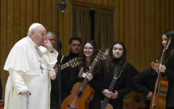 Pope Francis uses a cane and puts his hand to his ear as he stands in front of a group of young guitar players