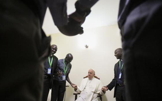 Pope Francis, sitting, holds hands with African Jesuits standing in a circle