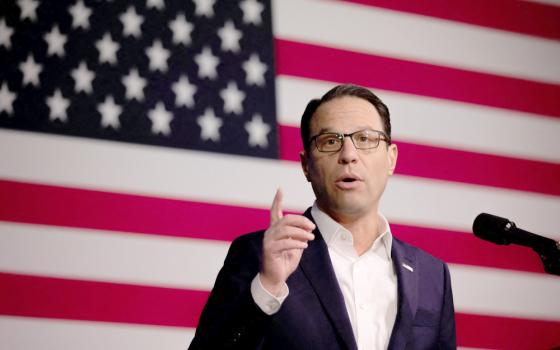 A white man with glasses stands behind a microphone and in front of an American flag
