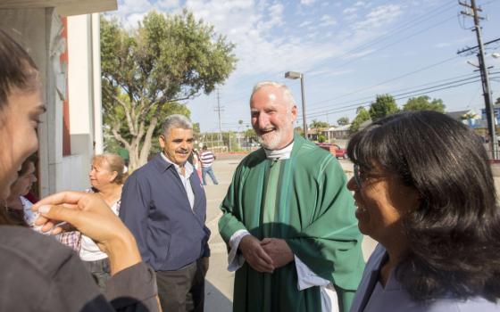 A white man with white hair wears green vestments and talks to a group of people outside