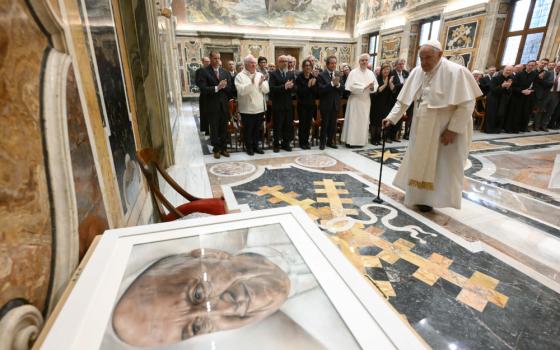 Pope Francis stands with a cane and looks at a portrait of him that is much bigger than him with an audience in the background