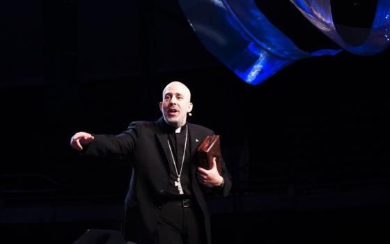 A bald man in a black cassock and a pectoral cross gestures energetically on a dark stage