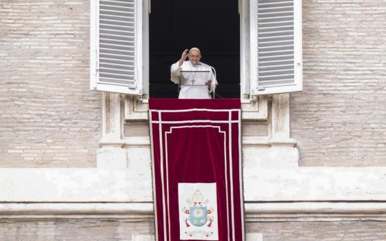 Pope Francis raises his hand while standing in a window behind a clear podium