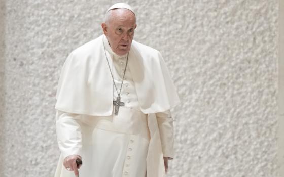 Pope Francis walks with a cane by a white wall