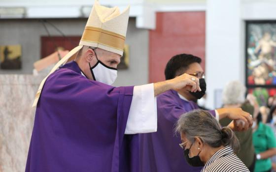A man wearing a mitre, a mask, and purple robes sprinkles ashes over the head of a woman wearing a mask