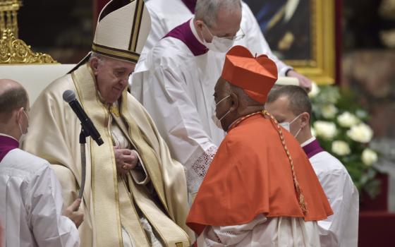 Cardinal Gregory, wearing a mask and a red biretta, kneels in front of Pope Francis, wearing a mitre