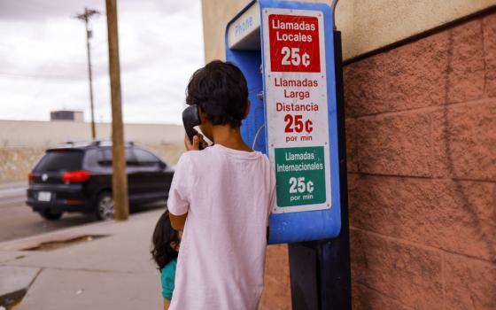 A brown-skinned child with short hair has their back to the camera and a payphone to their ear