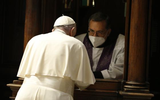 Pope Francis leans towards a masked priest wearing white and purple in what almost looks like a window sill, a confessional
