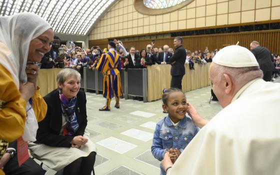 Pope Francis touches the cheek of a smiling Black girl. A short-haired white woman and Brown woman with her hair covered smile as they watch.