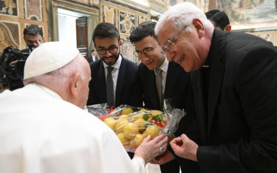 A group of men in suits and cassocks gather around Pope Francis and hand him a seran-wrapped container of lemon-looking fruit