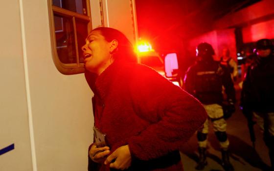 A woman leans on an ambulance in severe distress, clutching a cellphone, in red emergency light