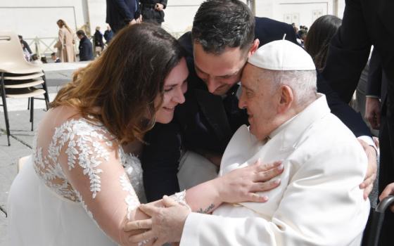 A woman in a wedding dress and a man put their heads close to Pope Francis, who is sitting in a wheelchair