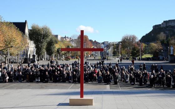 A large group of people dressed in black kneel in front of a large red cross outside