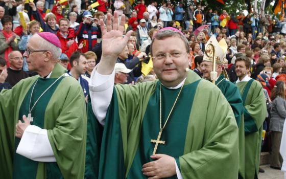 A white man wears glasses, a green robe, a violet zucchetto, and a pectoral cross and waves at a parade audience