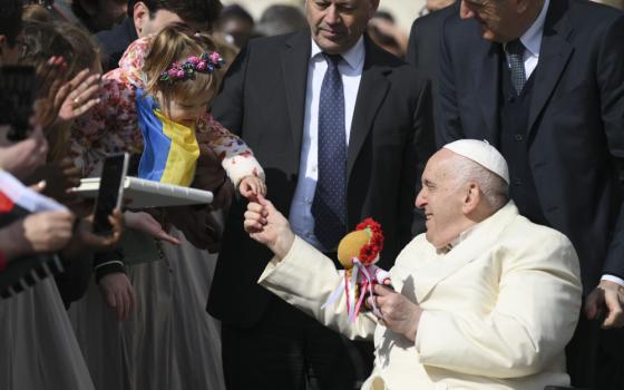 Pope Francis holds a little girl's hand. The little girl is wearing a Ukrainian flag. Pope Francis holds a doll wearing a flower crown.