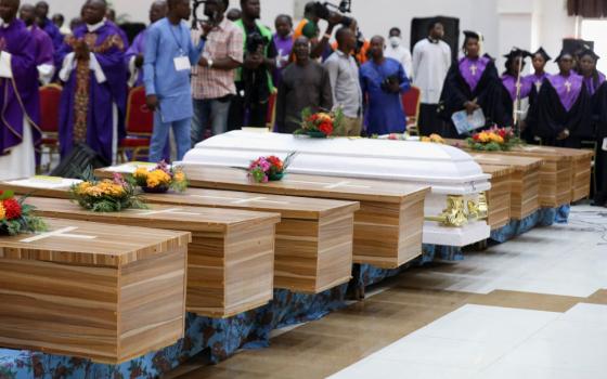 A row of caskets have flowers placed on them. They are surrounded by people in various church garments and other people with news cameras.