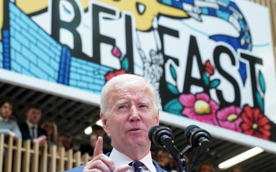 Joe Biden speaks into microphones in front of a mural that says "Belfast"