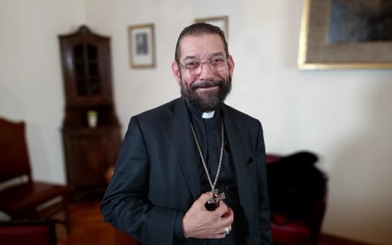 A bearded black-haired man wearing a black cassock, a black jacket, and pectoral cross smiles at the camera