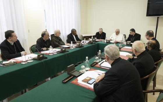 A group of men in black cassocks sits at long tables in a U shape with Pope Francis, without his zucchetto, at the head