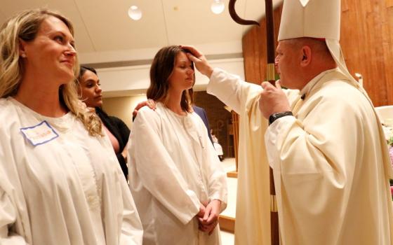Bishop makes sign of cross on forehead of young woman, while another woman looks on.