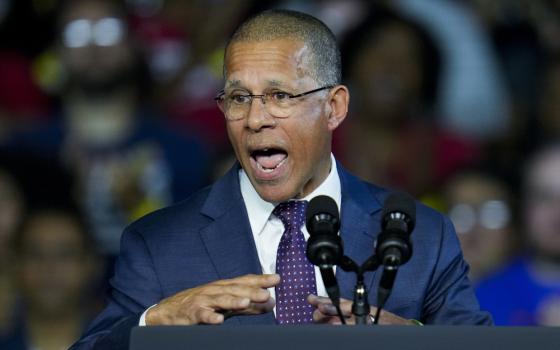 A light-skinned Black man wearing glasses and a suit speaks into a microphone at a political rally