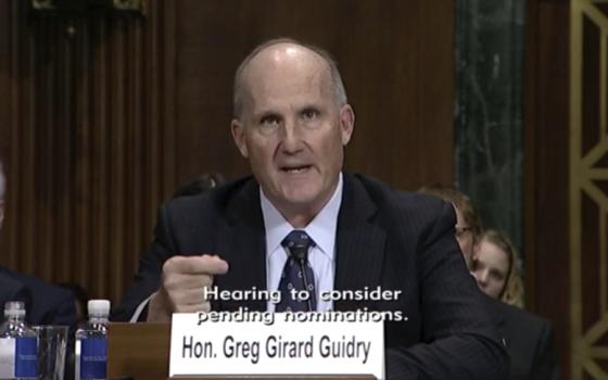 A bald white man in a suit sits behind a paper placard that reads "Hon. Greg Girard Guidry"