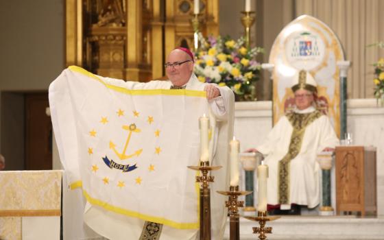 A white man with an amaranth zucchetto holds a Rhode Island state flag in front of a cathedral. A white man with a mitre is visible in the background.