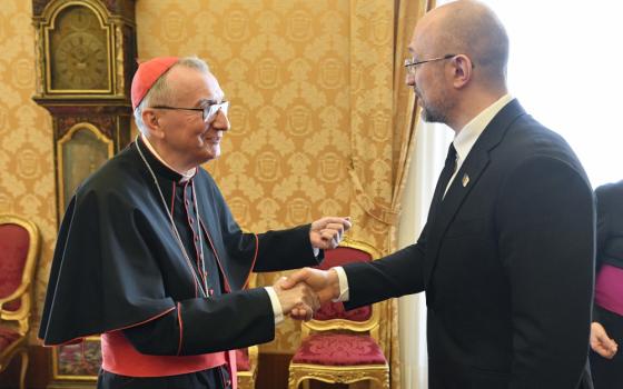 A man wears a cardinal's cassock and zucchetto and shakes a bald man in a suit's hand