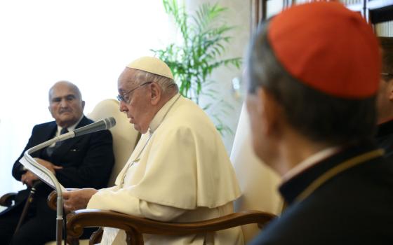 Pope Francis speaks into a microphone with a man in a suit and a man in a red zucchetto looking on