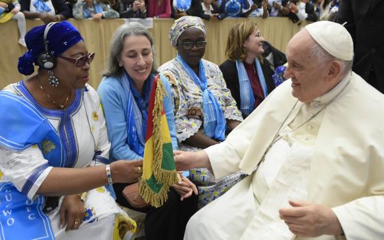 Pope Francis sits in front of a row of women dressed in blue and reaches out to touch a red, yellow and green flag one woman carries