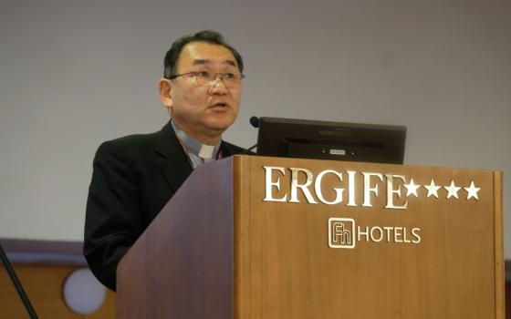 An East Asian man with a clerical collar speaks behind a podium labelled "Ergife Hotels"