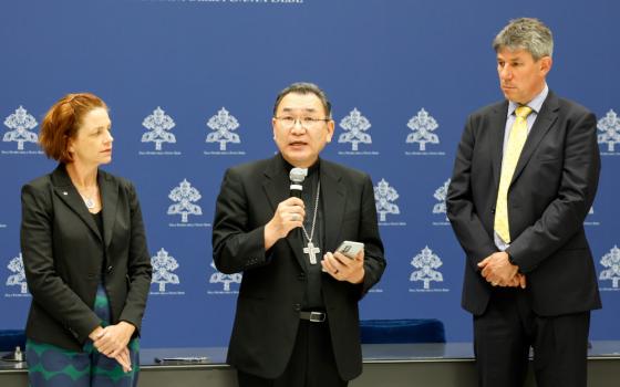 A man wearing a black suit, clerical collar, pectoral collar and glasses speaks into a microphone while a man and women in professional clothes stand on either side