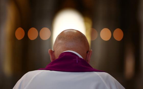 A bald person is seen from behind wearing white and a stole and bowing their head 