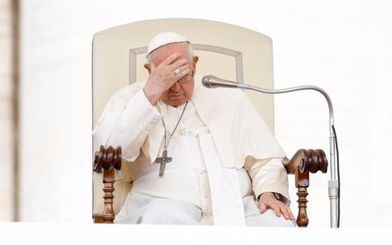 Pope Francis looks down and places his head on his forehead as he sits behind a microphone