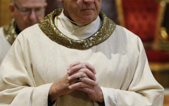 A white man with glasses wears a white and gold chasuble and folds his hands in front of him