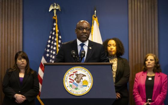 A bald Black man speaks behind a podium with an Illinois seal into several microphones, as flags and others stand behind him