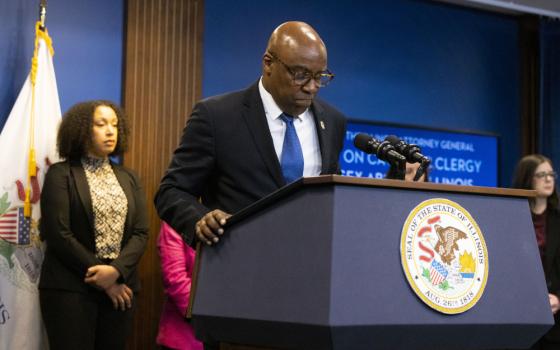 a Black bald man with glasses and a suit looks down at a podium with microphones