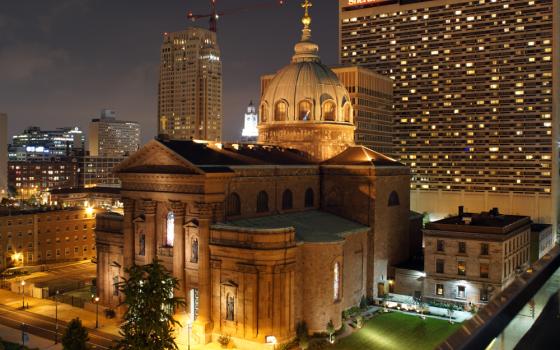 A cathedral with a dome and columns is lit up at might with tall buildings surrounding it