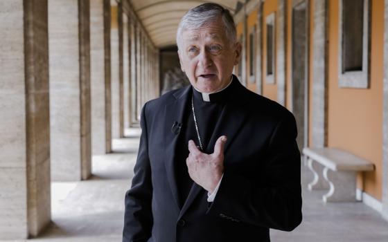 An older white man with blue eyes wears a clerical collar, black coat, pectoral cross, and microphone as he speaks to the camera