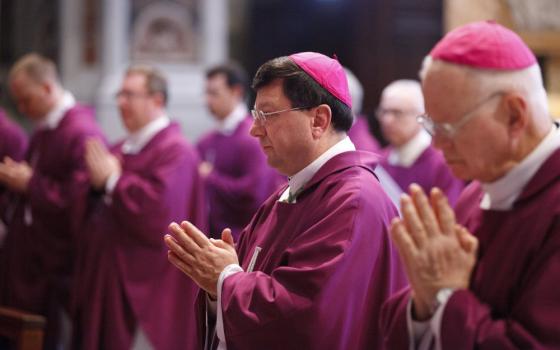 A brown-haired man wearing glasses and a violet zucchetto and vestments holds his hands together in prayer among many other bishops