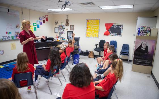A blonde woman in a red dress stands in front of a group of children in red uniforms in a music classroom.