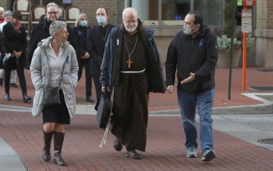 An older white man wearing a brown Franciscan habit walks between laity and priests. Some where face masks.