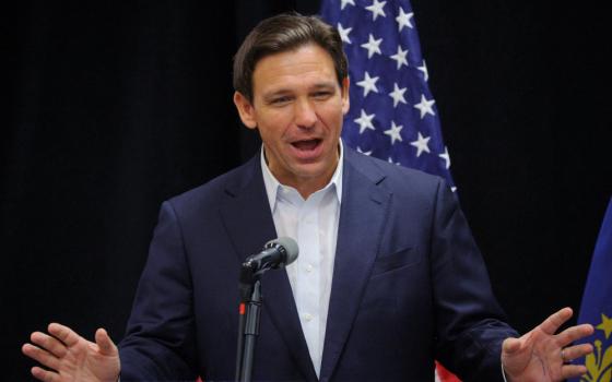 Ron Desantis wears a suit jacket with no tie and speaks into a microphone while standing in front of an American flag