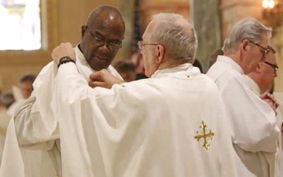 An older white man wearing glasses and a white chasuble adjusts the vestments of a bald Black man