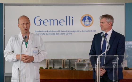 A white man in a white doctor's coat with scrubs peaking through stands next to a white man in a blue suit and tie. Both use microphones in front of a banner that says Gemelli