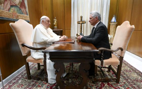 Pope Francis sits in a chair on one side of the table as an older white-appearing man sits in an identical chair on the opposite side