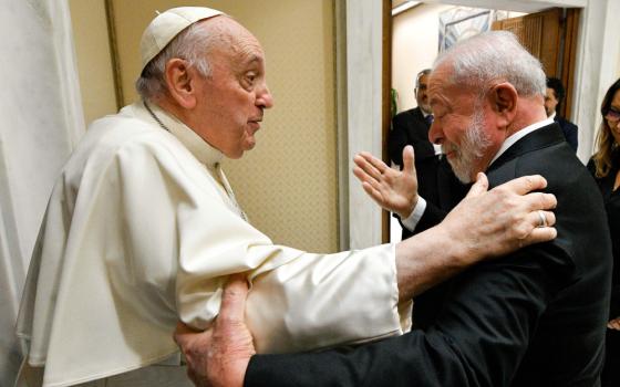 Pope Francis grips the shoulder of an older, white-appearing man in a suit, who in turn grips Pope Francis' upper arm